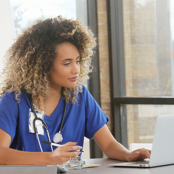 A nurse studying on a laptop while taking handwritten notes