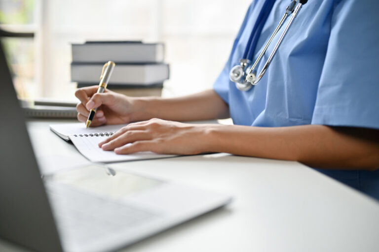 close up of a nurse writing notes while working on laptop