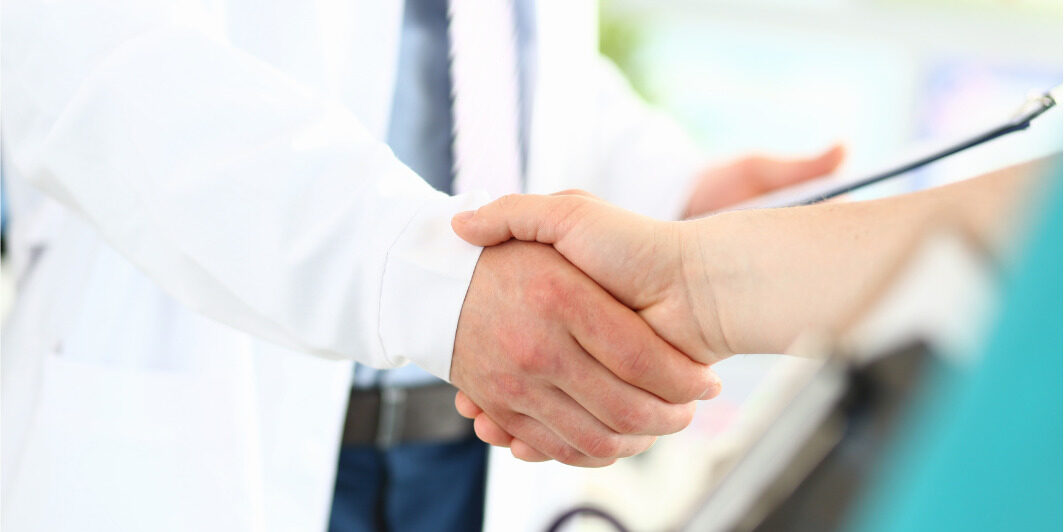 Close up of two people shaking hands, one wearing a white lab coat and tie while holding a clipboard.