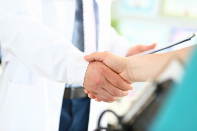 Close up of two people shaking hands, one wearing a white lab coat and tie while holding a clipboard.